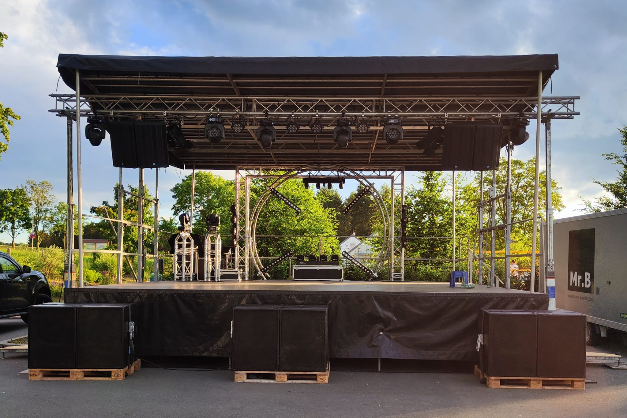 Bühnensystem vor Hecken und Bäumen, Bühne mit Belichtung am oberen Rigging und radialer Beleuchtung hinter den Musikern
